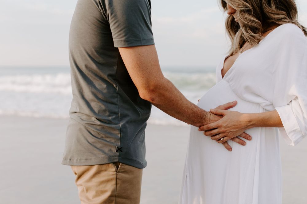 Person in a grey shirt and khakis caressing a baby bump on the beach.