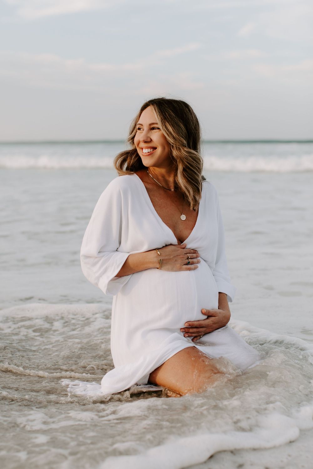 Person in flowy white maternity dress smiles while kneeling in ocean waves.