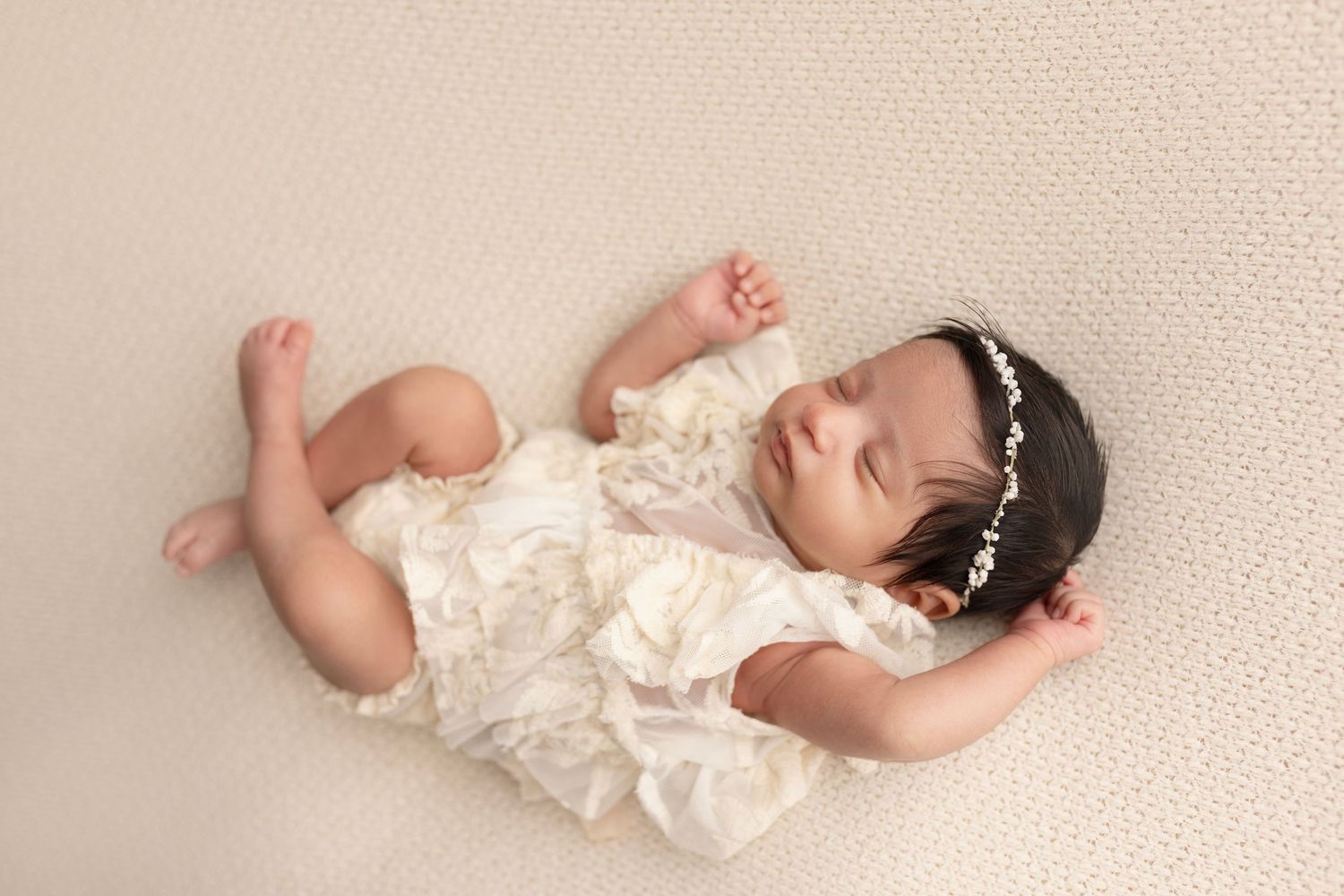 A newborn lays relaxed and tiny on a soft blanket. She wears a dainty headband and a white dress. Newborn photography Surrey Elizabeth Lang