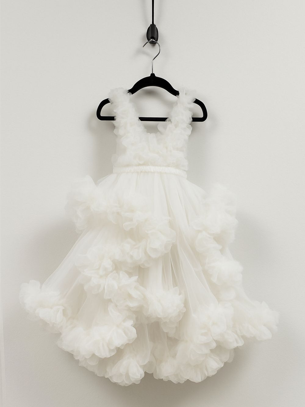 A white ruffled tulle flower girl dress with voluminous skirt hangs on a black hanger against a light wall.