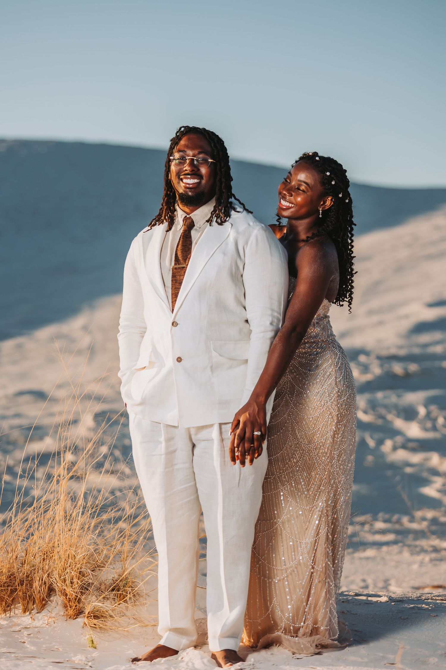 A romantic couple embraces on a sandy beach with dried grass and mountains in the background during golden hour.