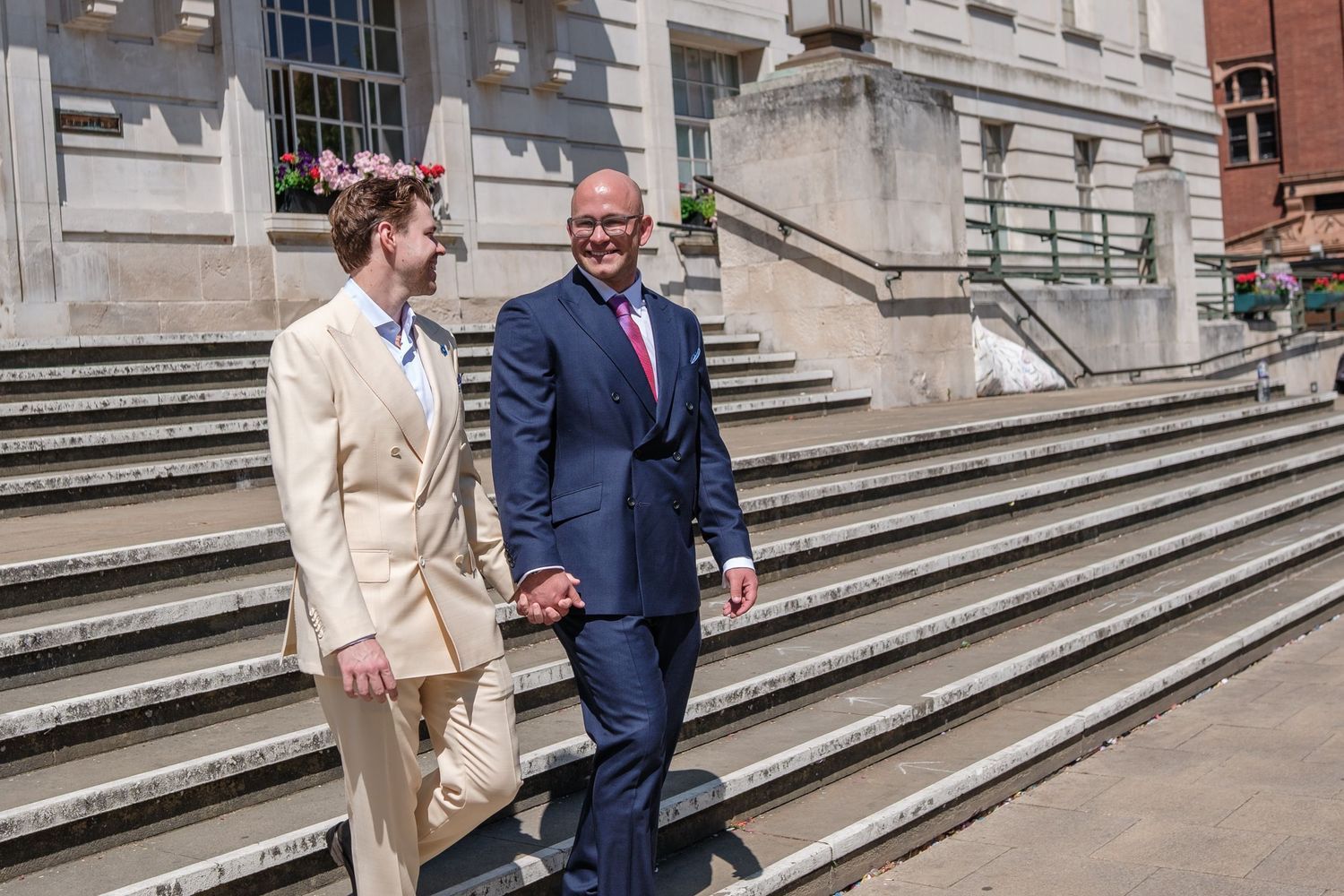 A couple in formal attire walks down stone steps outside a grand building, with the woman wearing beige and man in navy blue.