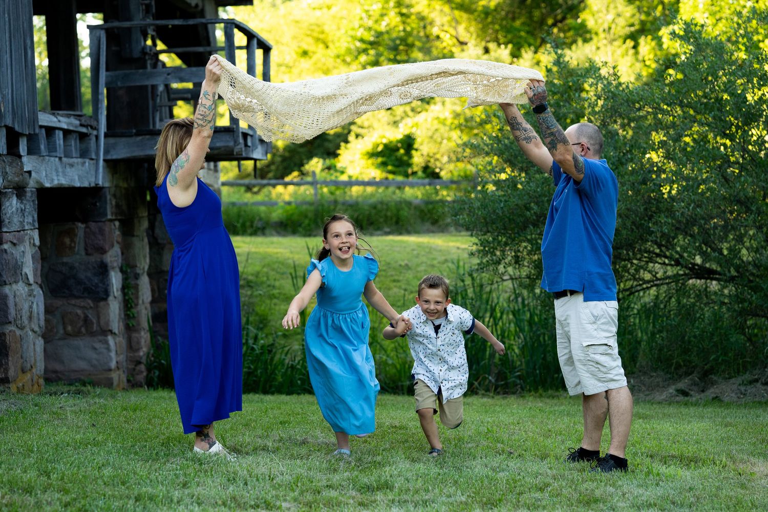 A group plays together outdoors on a sunny day, holding a white fabric aloft while running on green grass. Kids playing games during photos.