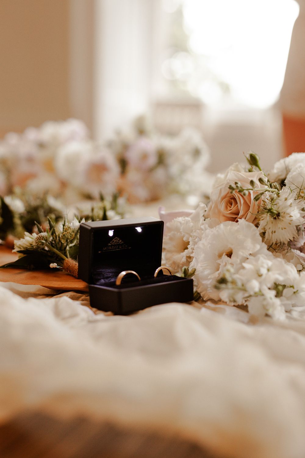 Wedding rings in black box surrounded by pink and white floral arrangements.