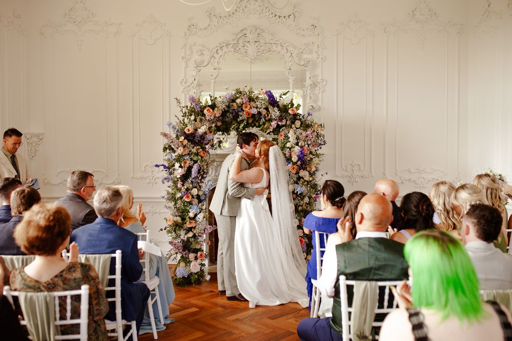 Wedding ceremony in an elegant white room with floral arch and seated guests.