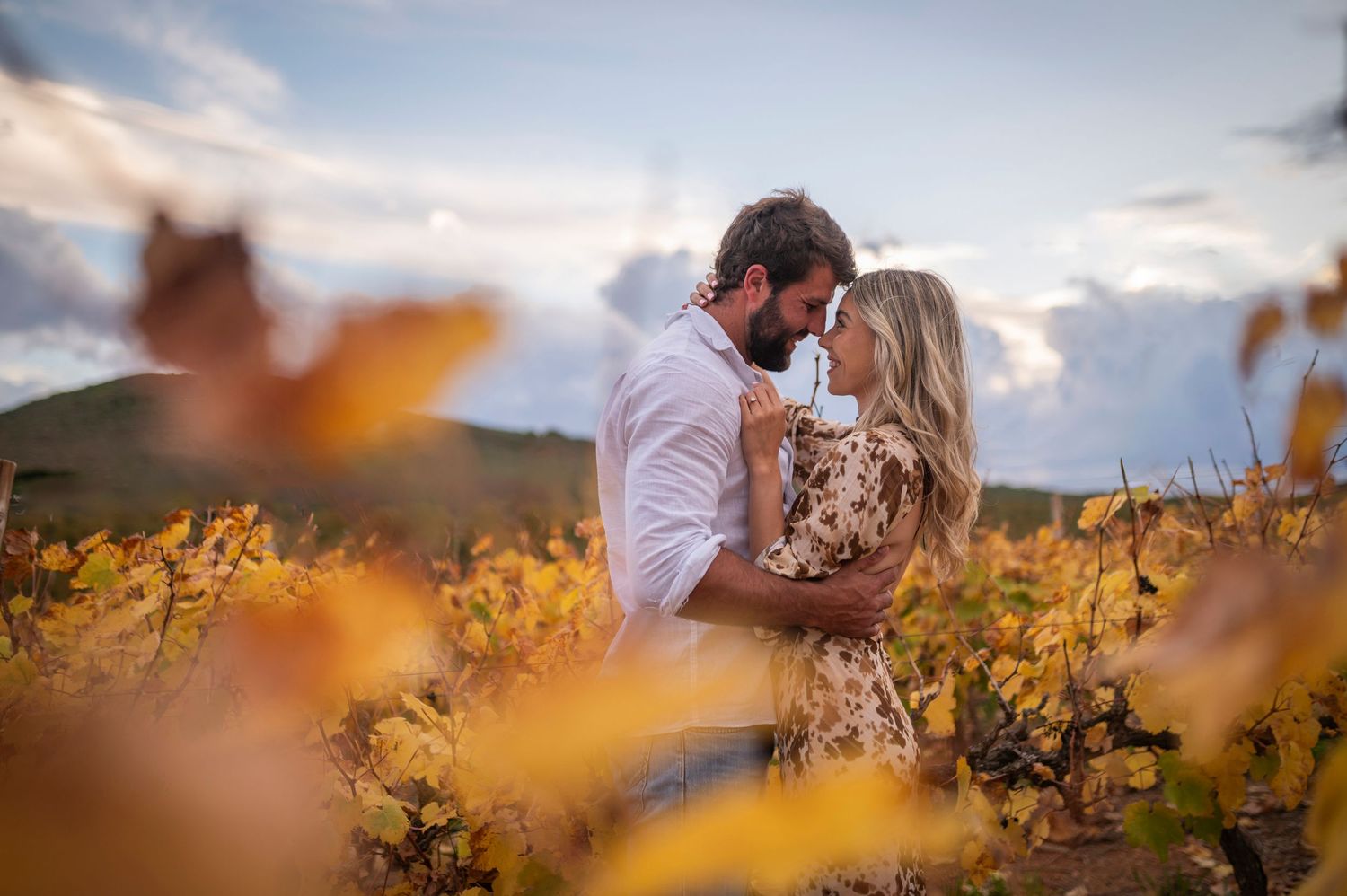A romantic couple embraces in a field of yellow wildflowers at sunset with mountains in the background.