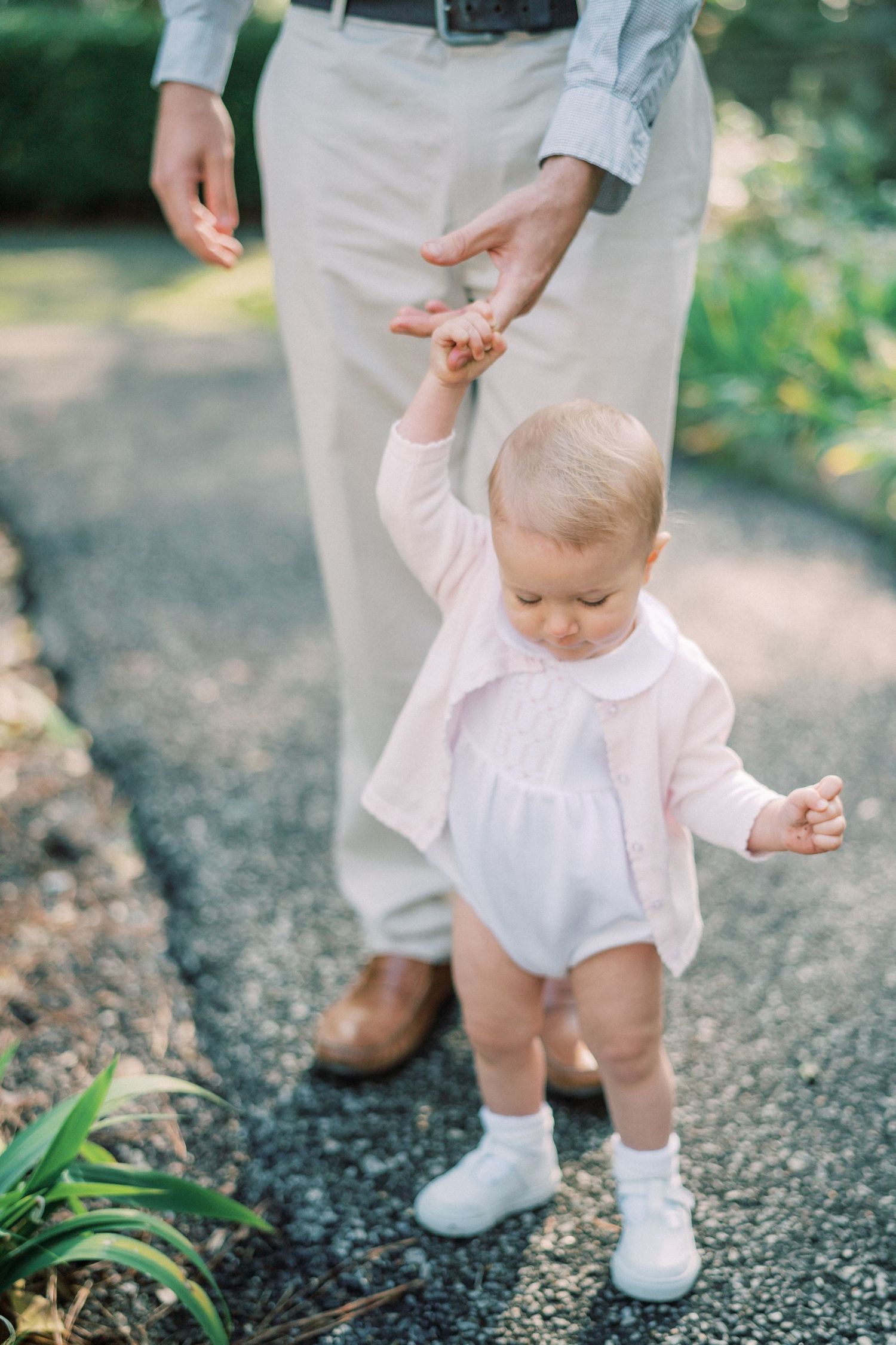 Small child in white romper taking first steps while holding parent's hand.