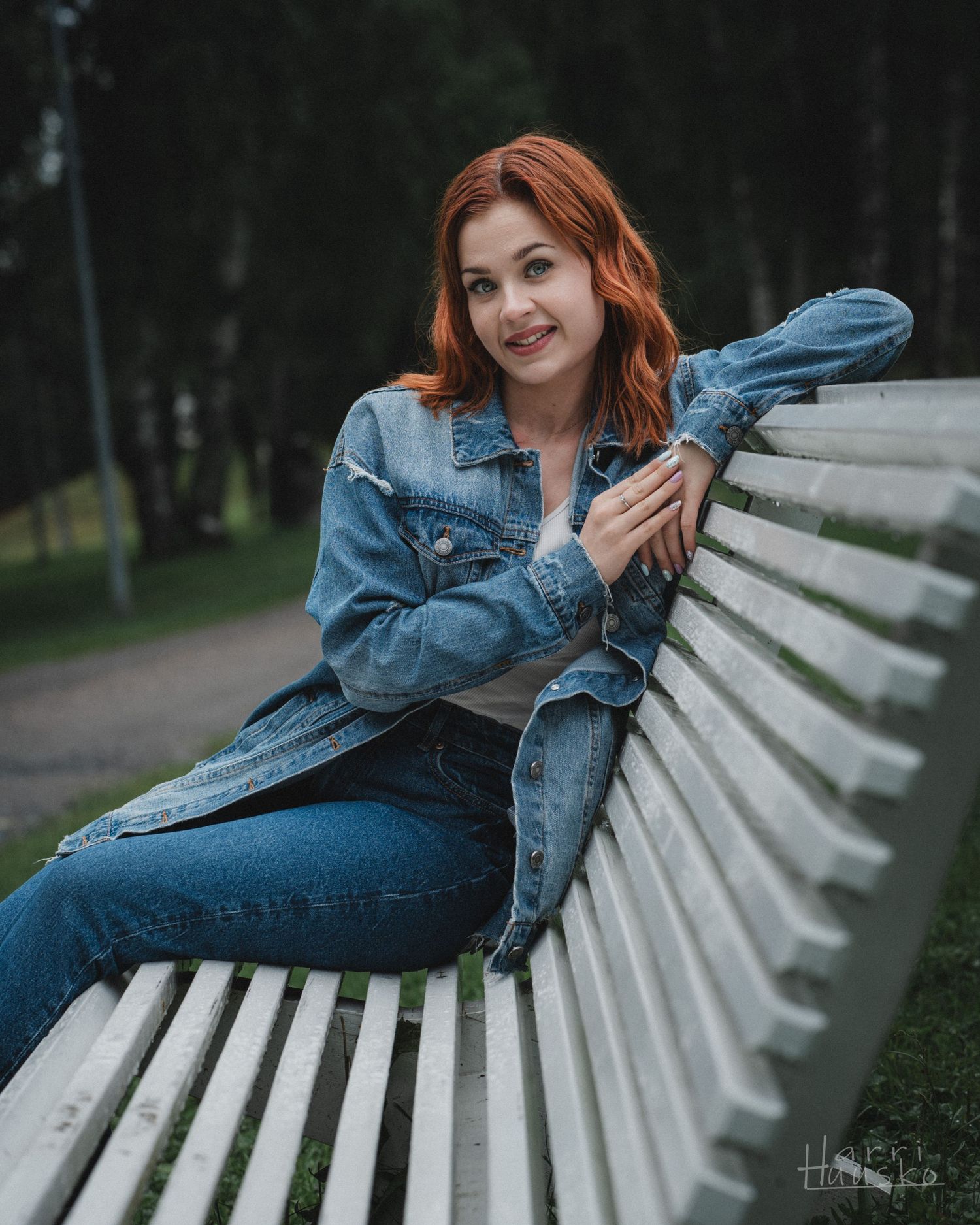 A casual denim-clad figure relaxes on a white wooden park bench surrounded by trees.