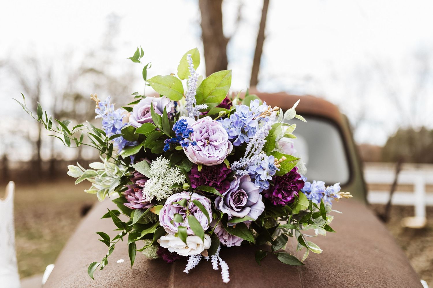 A romantic bouquet of purple roses and lilac flowers with green foliage arranged on a rustic brown surface outdoors.