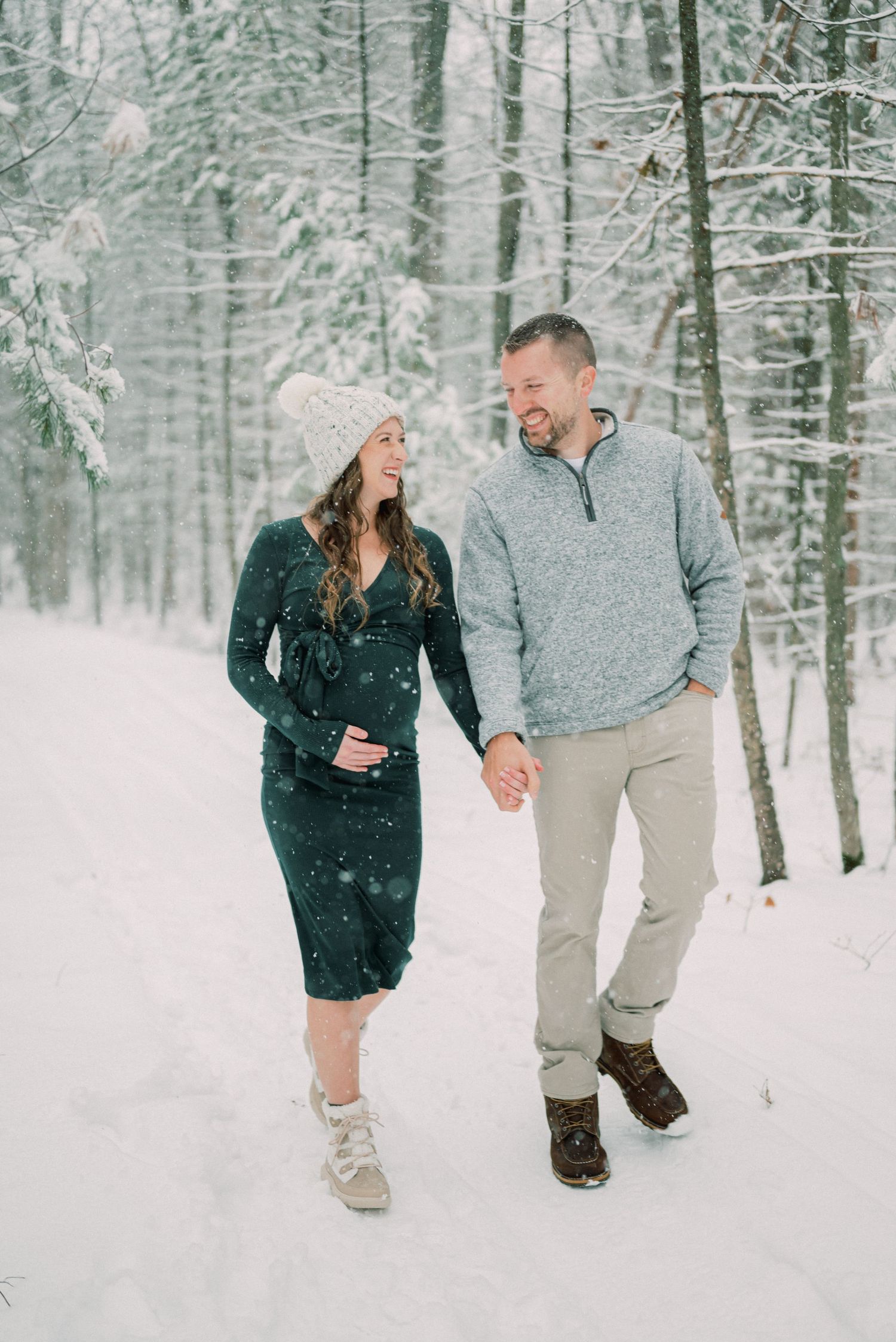 A winter maternity photo shoot with couple walking through snowy woods.