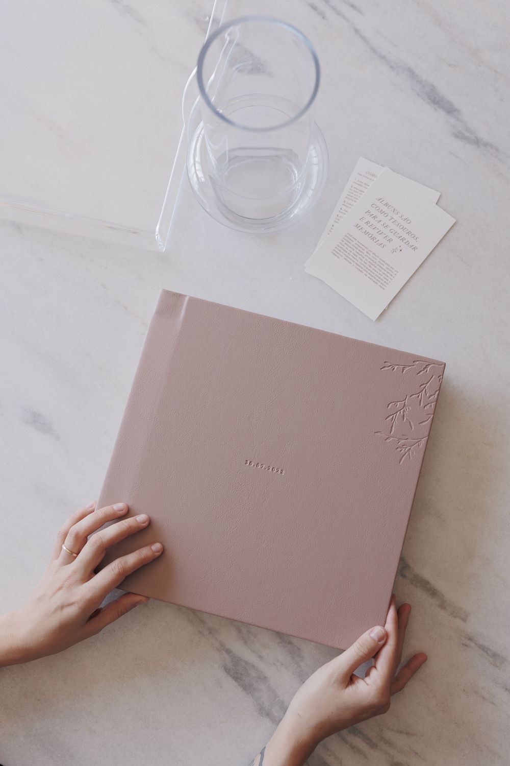 Hands holding pink leather photo album on marble surface with glass and cards.
