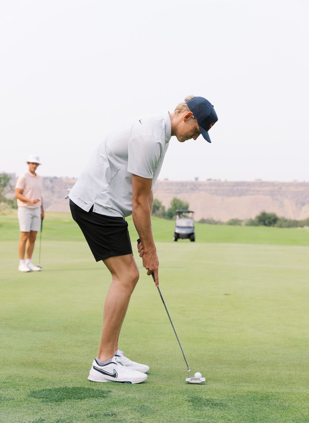 A golfer stands on a green course preparing to putt while another player and golf cart are visible in the background.