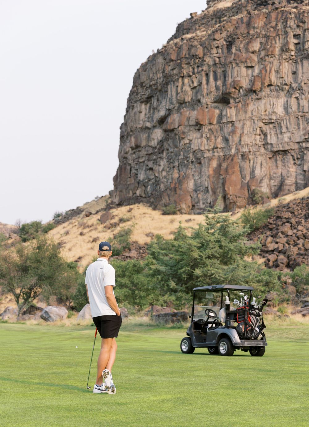 Golfer standing on green course with dramatic red cliffs and golf cart in background at desert golf resort.