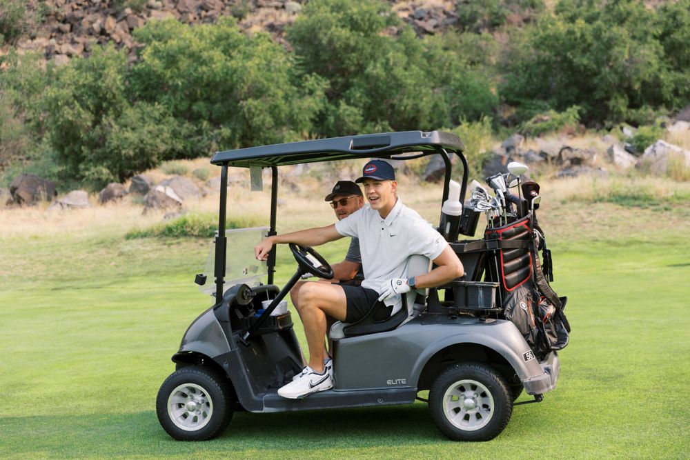 Two golfers riding in a gray golf cart on a scenic desert golf course with golf clubs visible in the back.