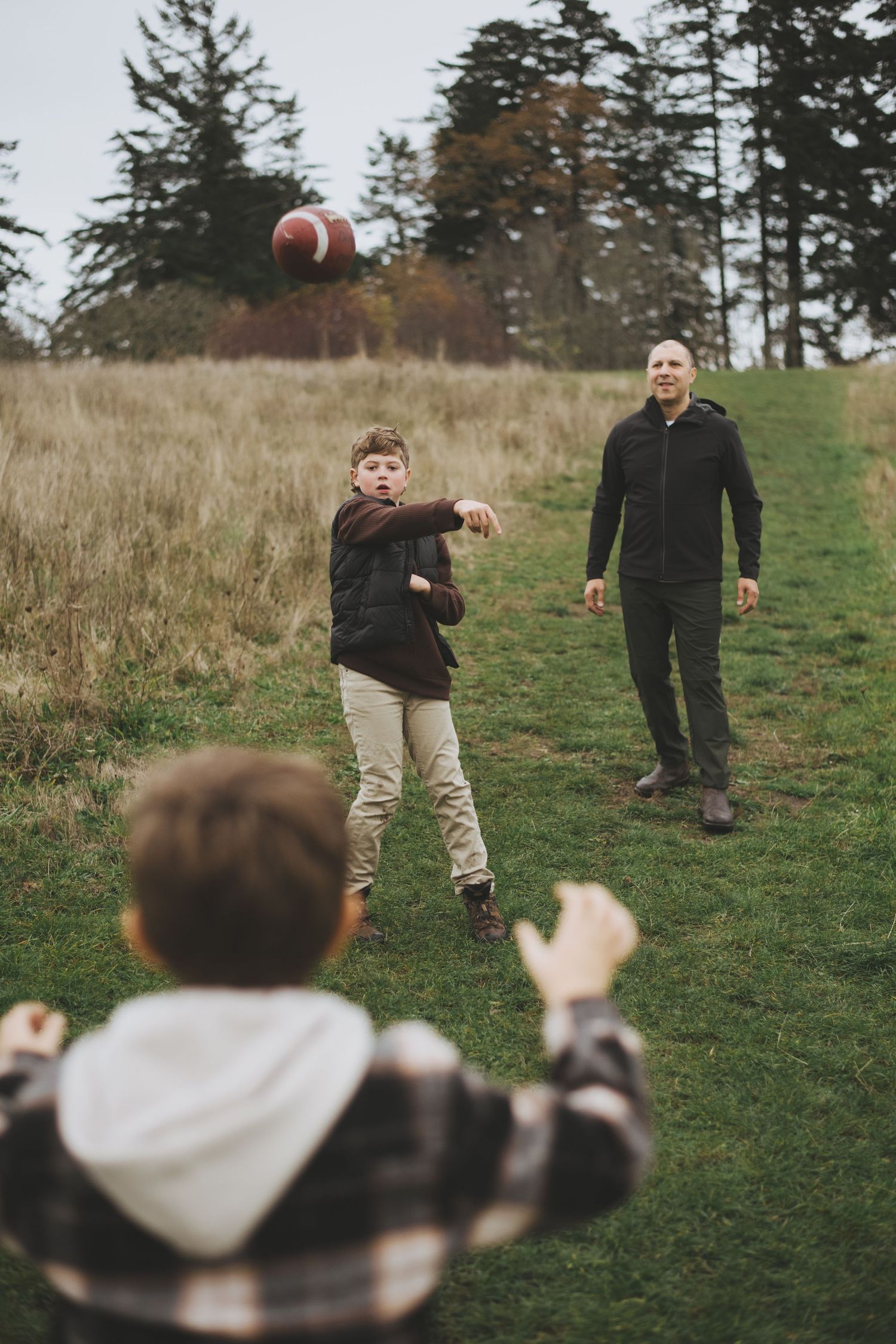 People playing catch with a football on a grassy hillside with evergreen trees in the background.