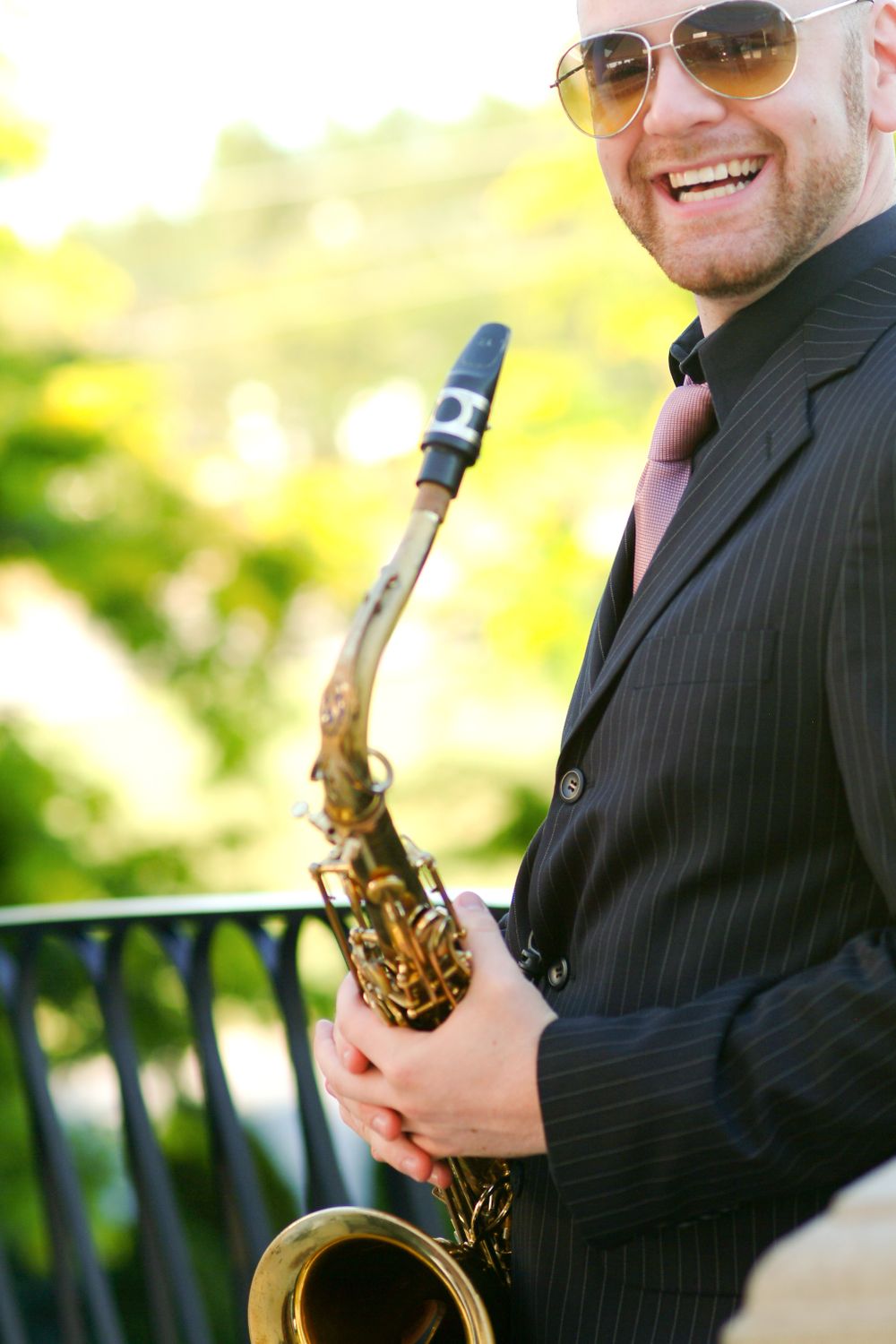 Musician in sunglasses and suit jacket smiling while holding a saxophone outdoors.