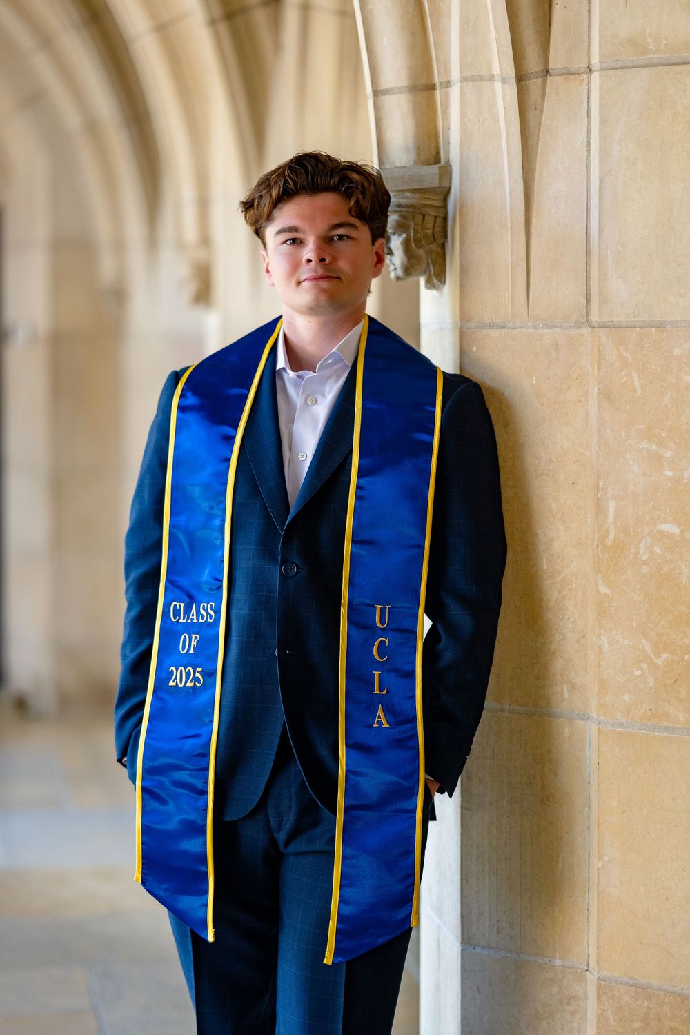 Graduate in navy suit wearing blue and gold UCLA graduation stole stands in arched hallway corridor.