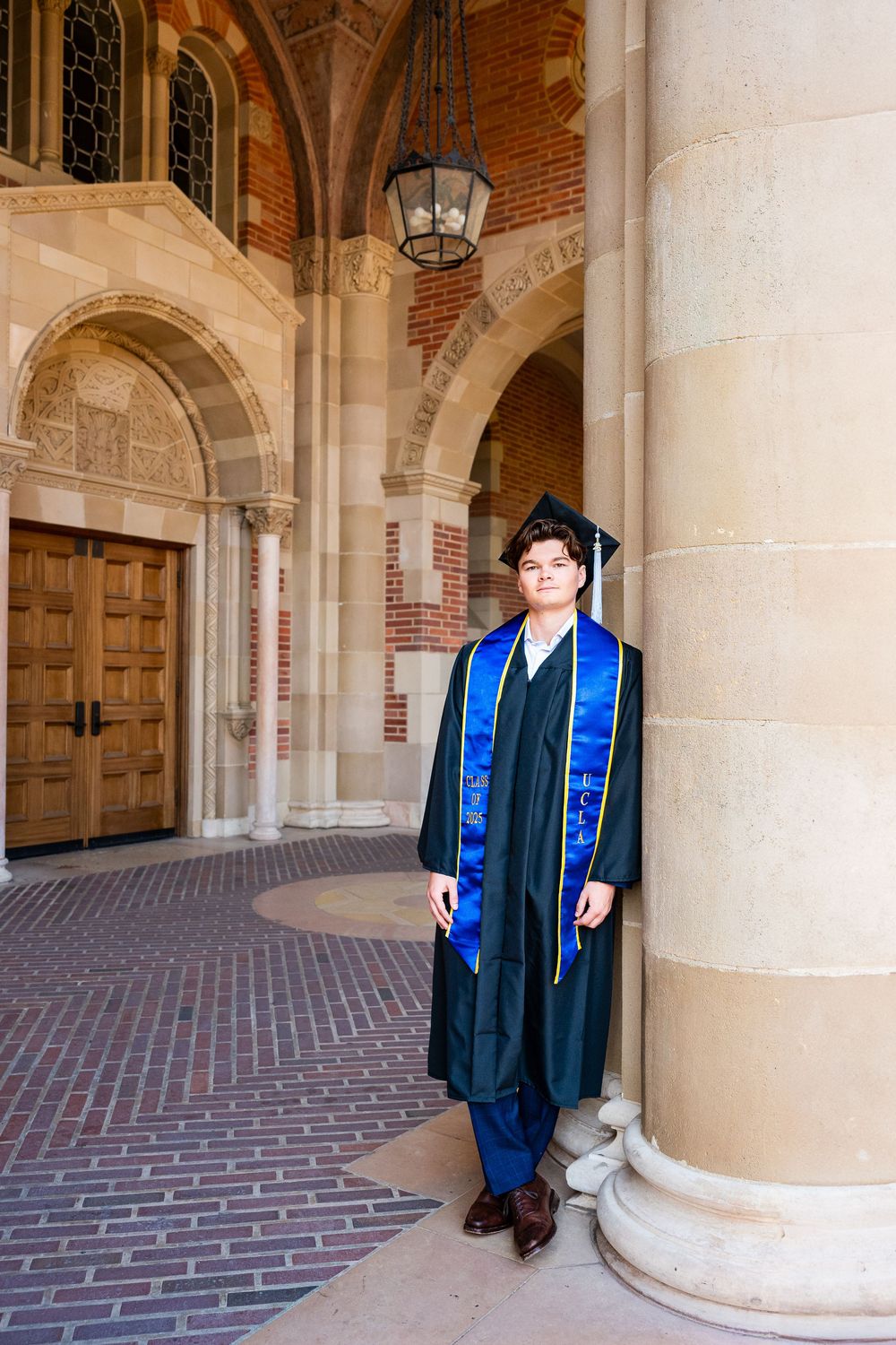 Academic graduate in cap and gown with blue sash stands in ornate brick and stone archway of university building.