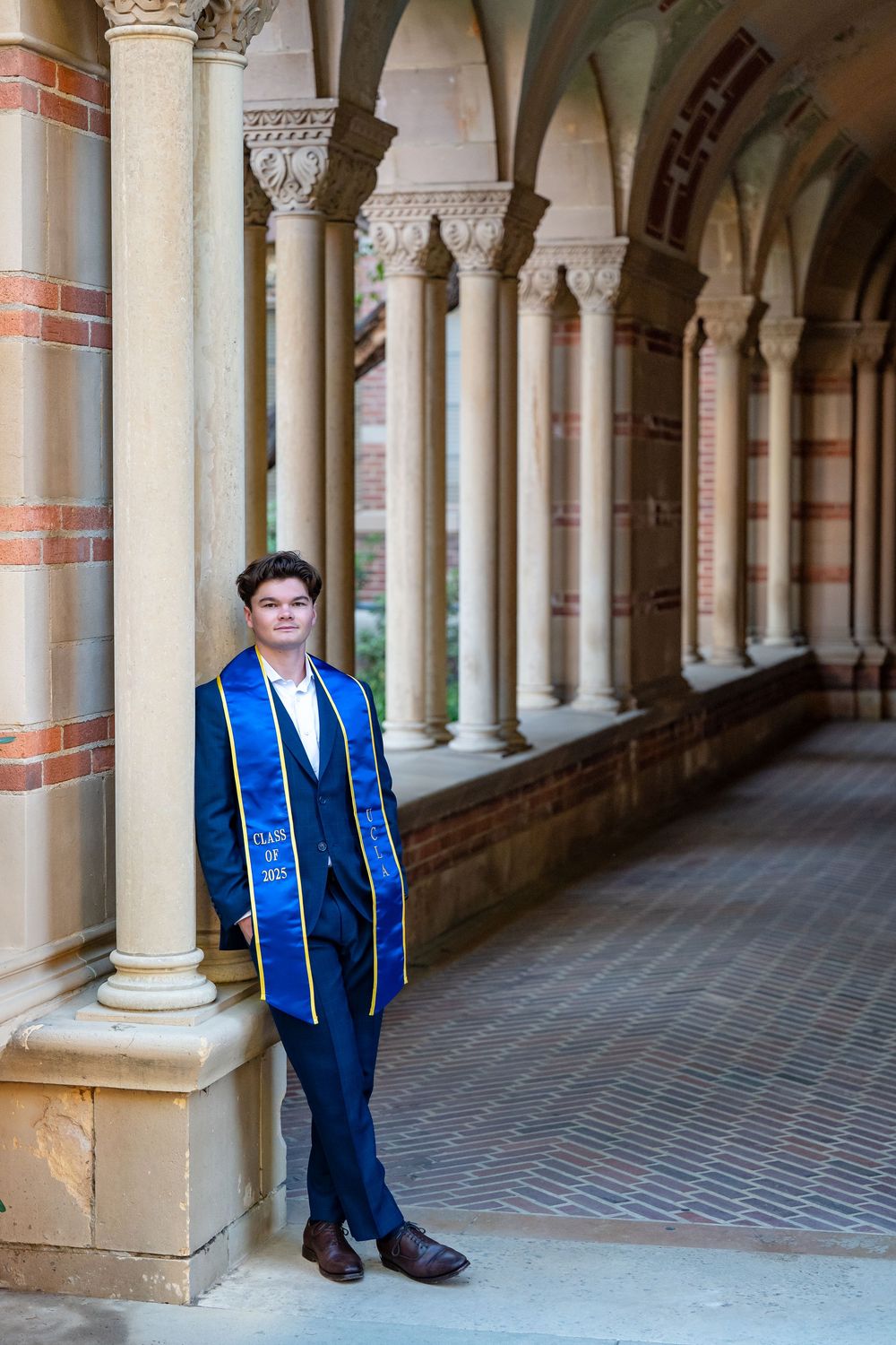 Graduate wearing blue sash and stole stands in ornate columned hallway of historic university building.