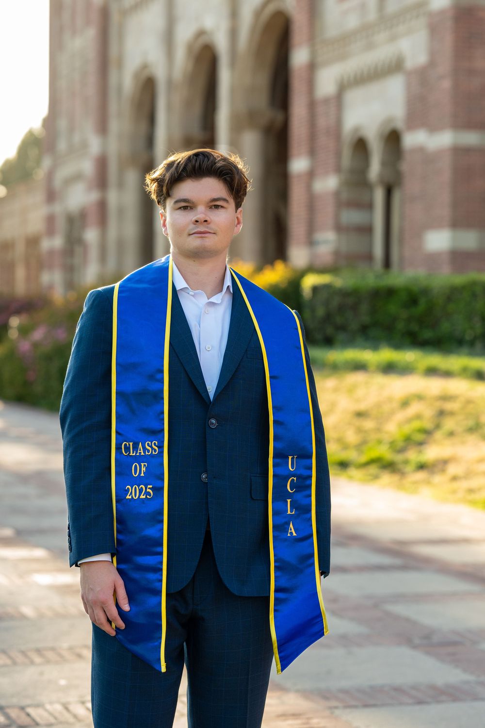Graduate wearing a blue and gold UCLA stole with class of 2025 sash stands in front of brick building exterior.