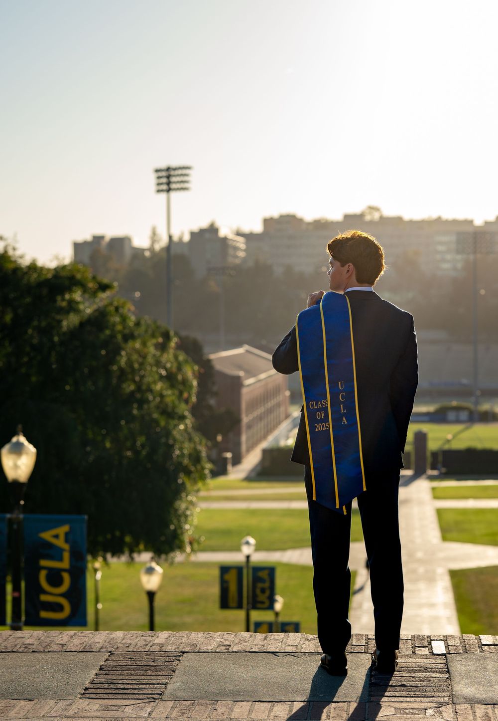 A graduate in formal attire stands on stairs overlooking the UCLA campus at sunset with their graduation stole.
