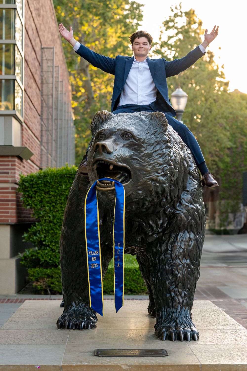 A celebratory pose atop the iconic UCLA Bruin Bear statue adorned with blue and gold ribbons on campus.