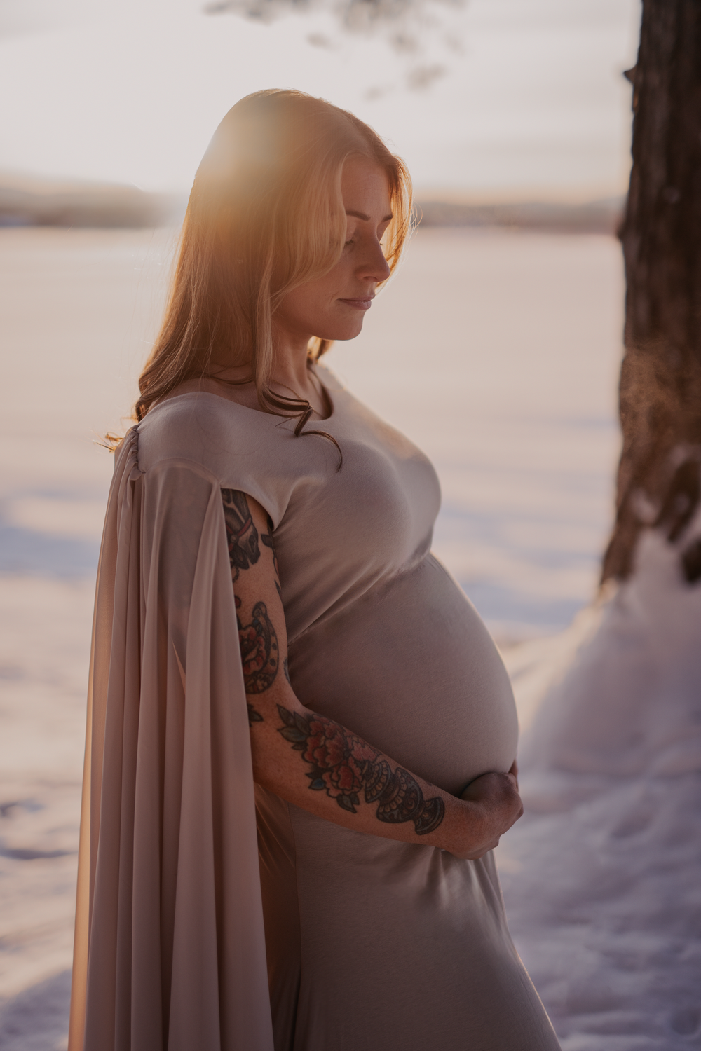 Serene winter maternity portrait with dramatic lighting and flowing cape.