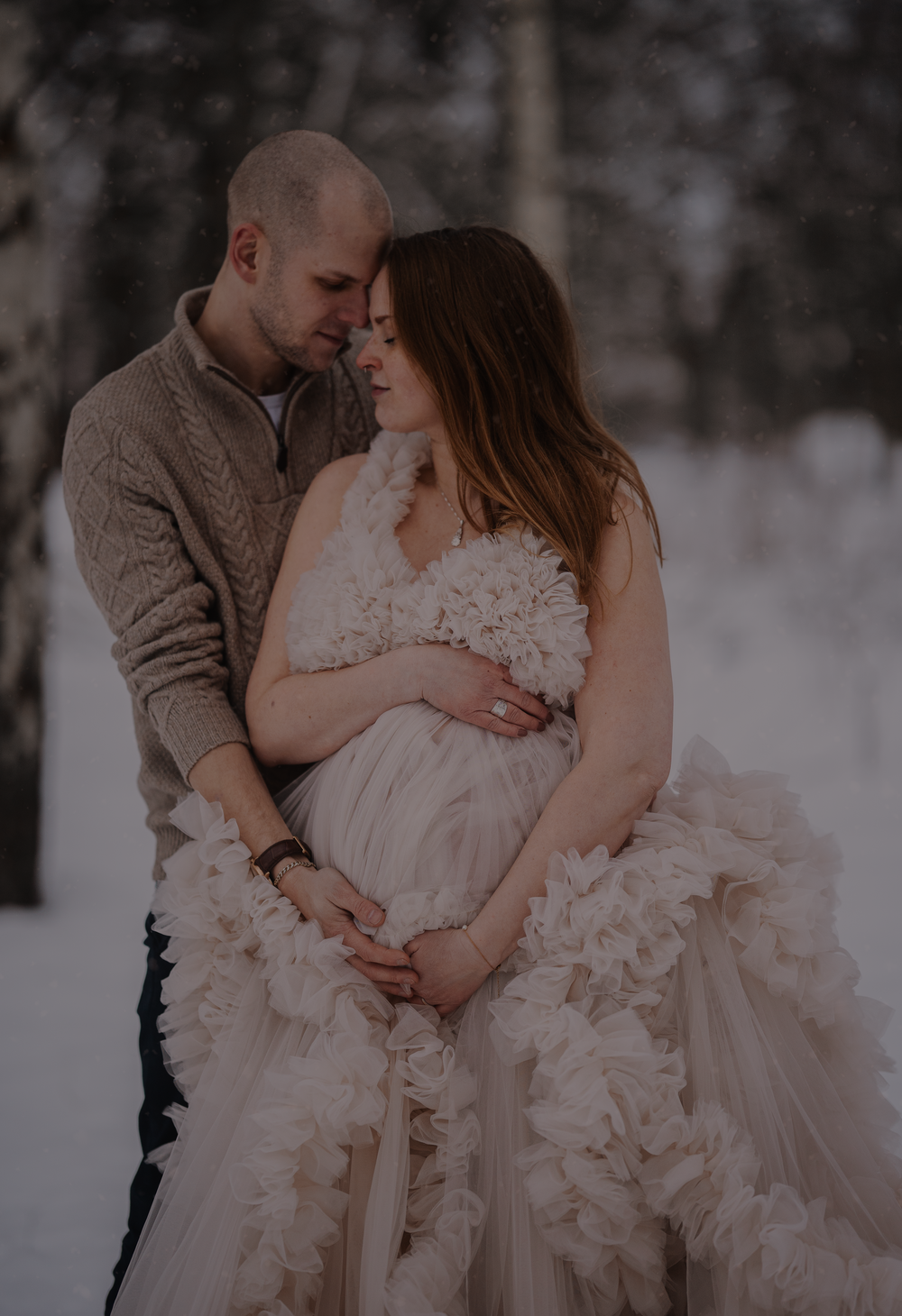 Romantic winter maternity portrait with dramatic tulle dress in snowy setting.