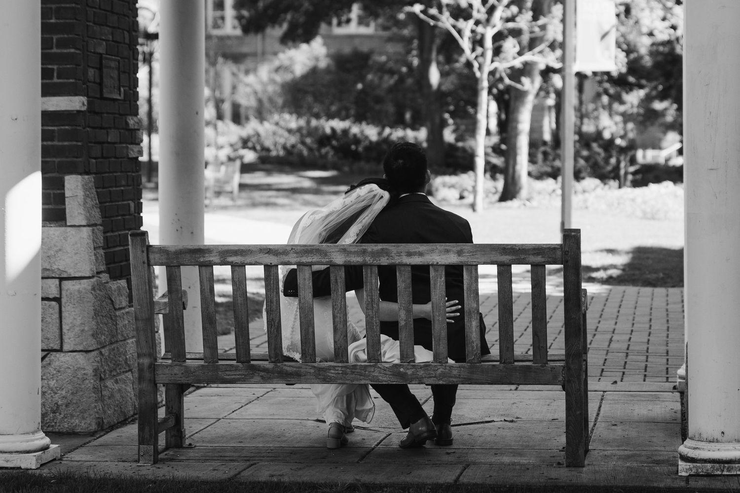 Black and white photo of a wooden bench between columns on a brick porch.