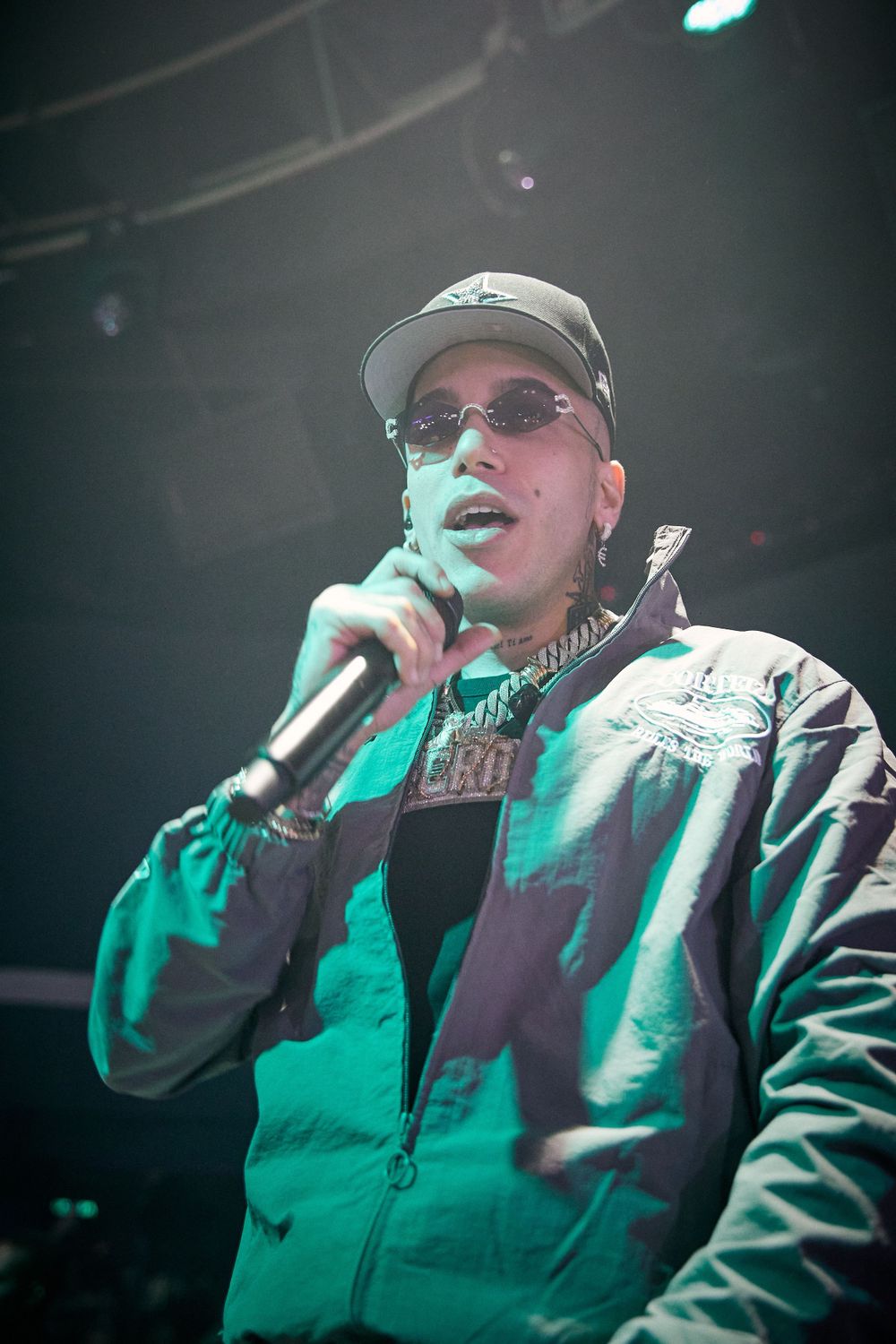 Performer in green jacket and baseball cap singing into microphone under stage lighting.