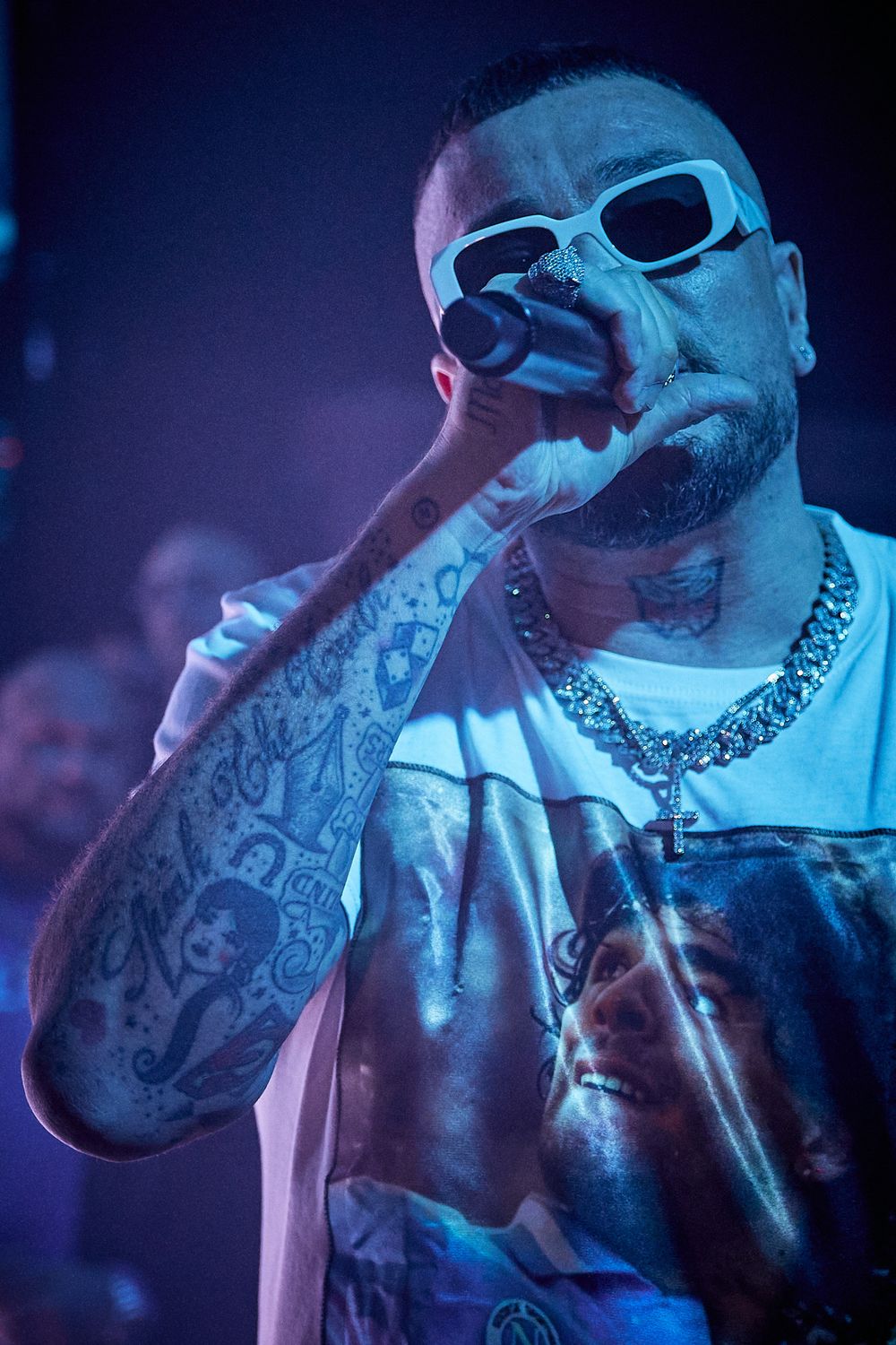Tattooed performer in white sunglasses singing into microphone under blue stage lighting.
