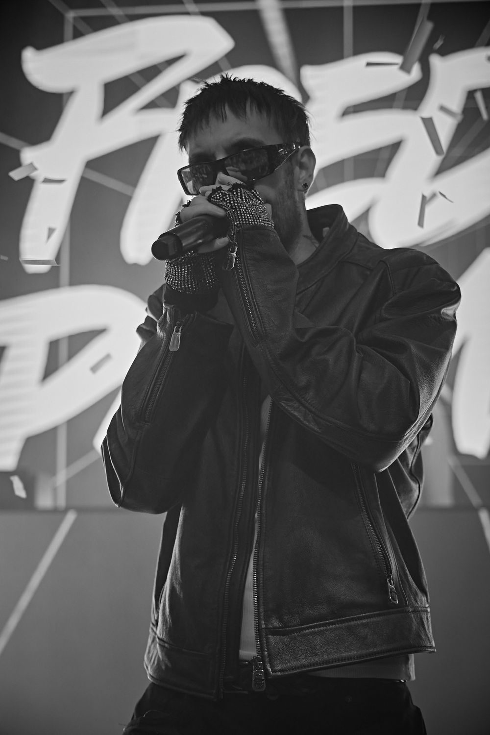 Black and white photo of performer in leather jacket singing into microphone against graffiti background.