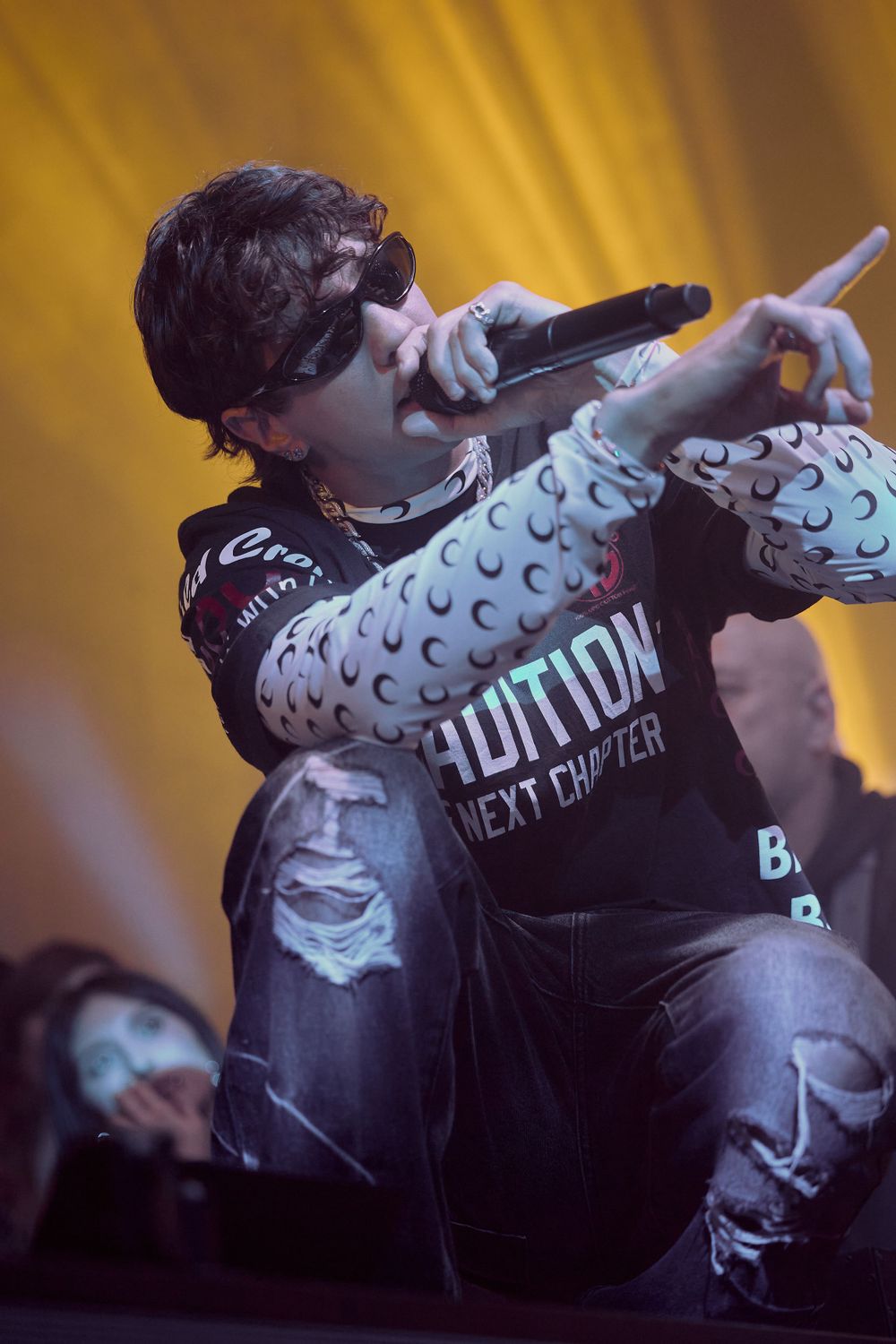 Performer in patterned shirt and sunglasses singing into microphone under yellow stage lighting.