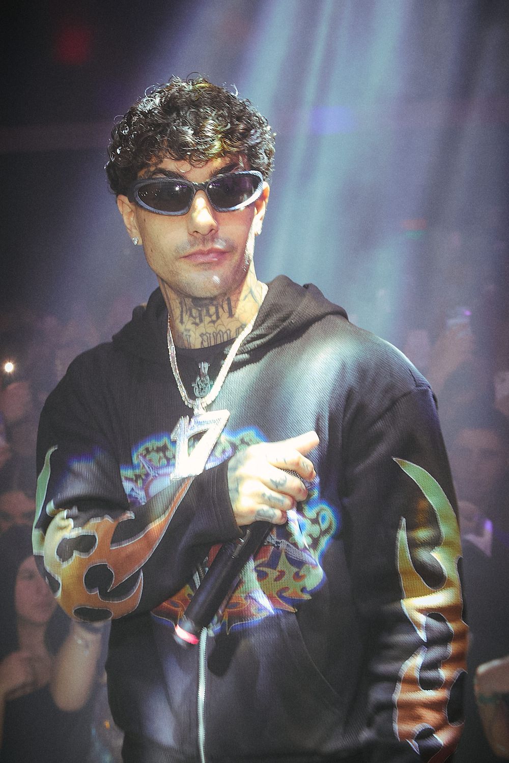 Artist performing on stage wearing black hoodie with graphic design and chain necklace.