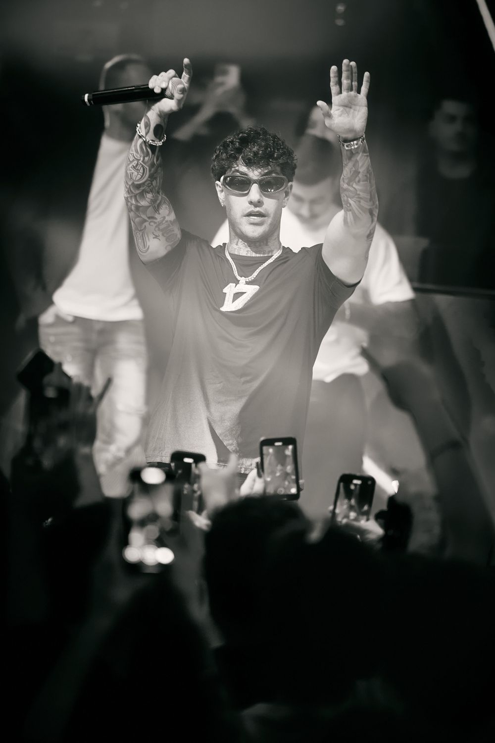 Performer wearing sunglasses raises hands during concert as crowd takes photos with phones.