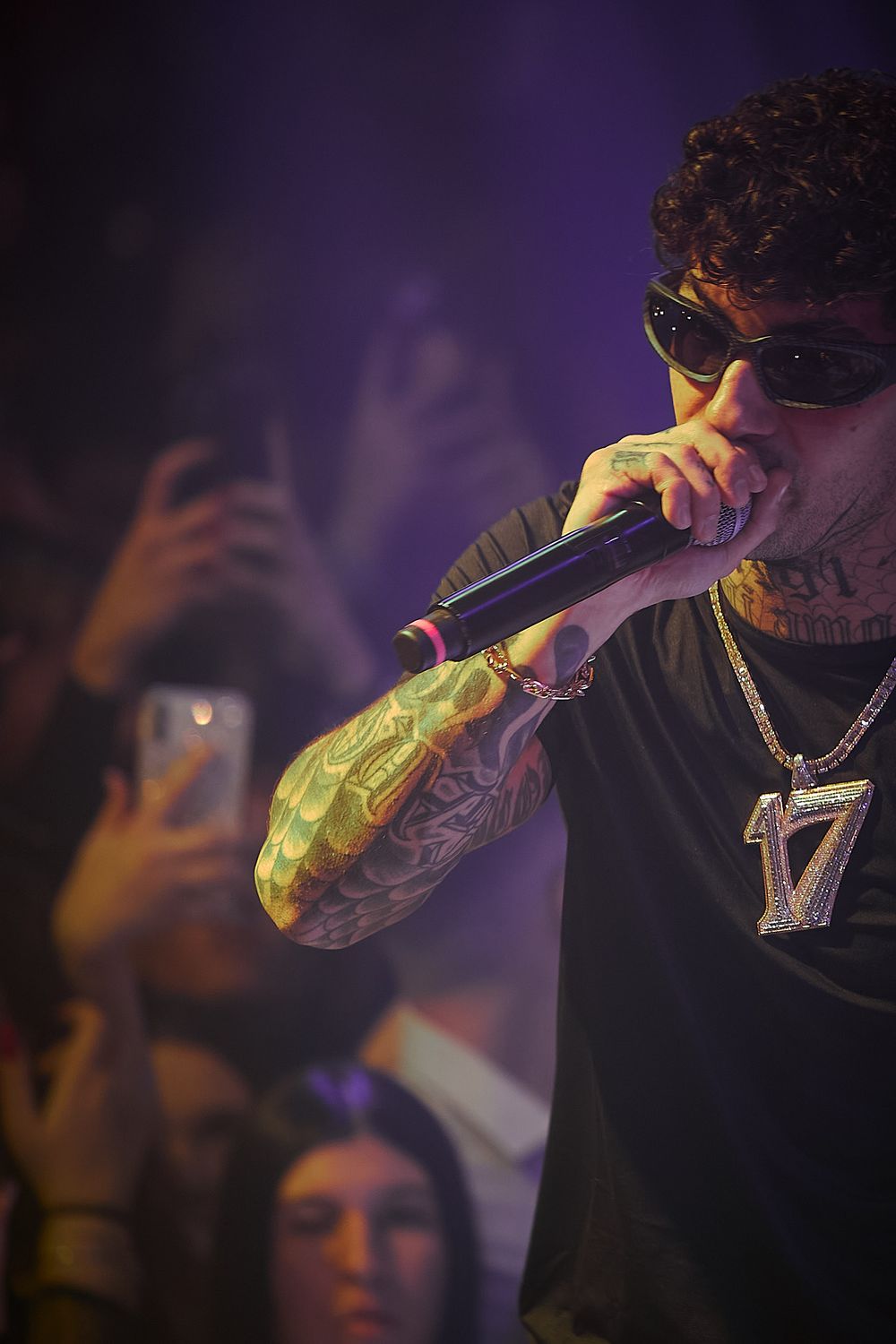 Performer with tattoos and sunglasses holds microphone during energetic concert performance.