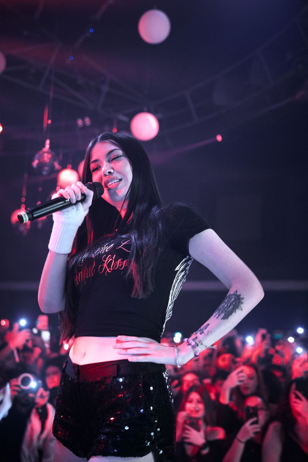 Concert performer in crop top and shorts energetically sings on stage with pink lighting.