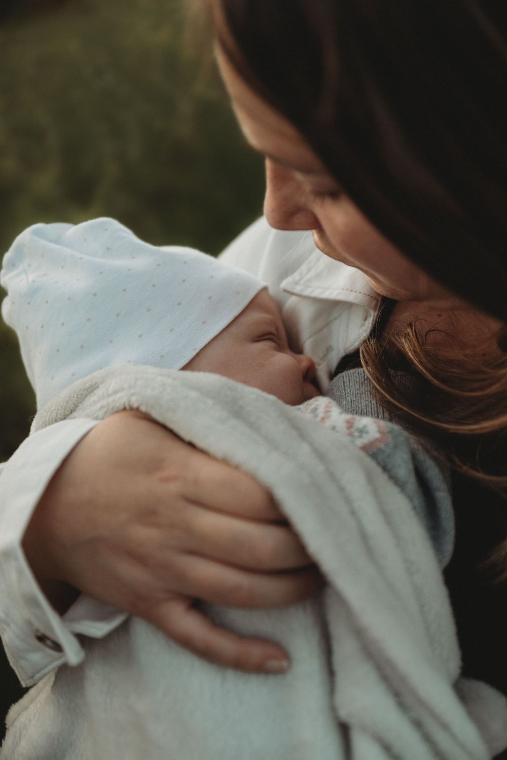 An intimate moment between parent and baby wrapped in a white blanket outdoors at sunset.