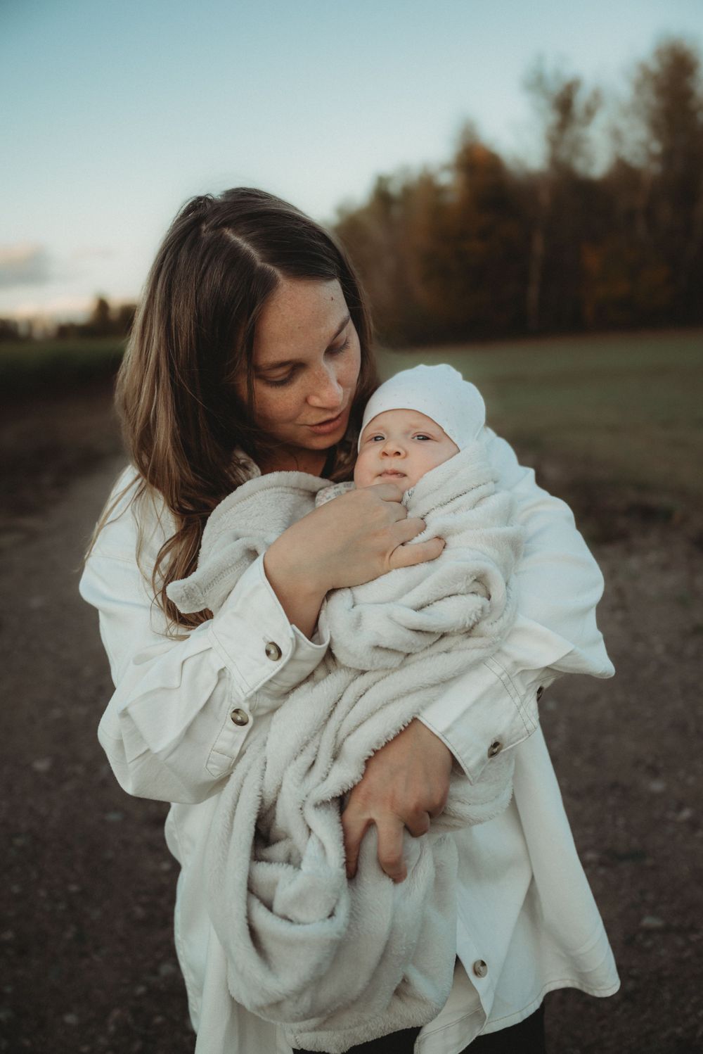 Parent holding infant wrapped in white blanket against autumn backdrop.