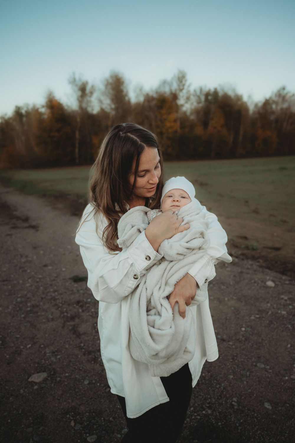 Parent embracing baby wrapped in white blanket on dirt path.