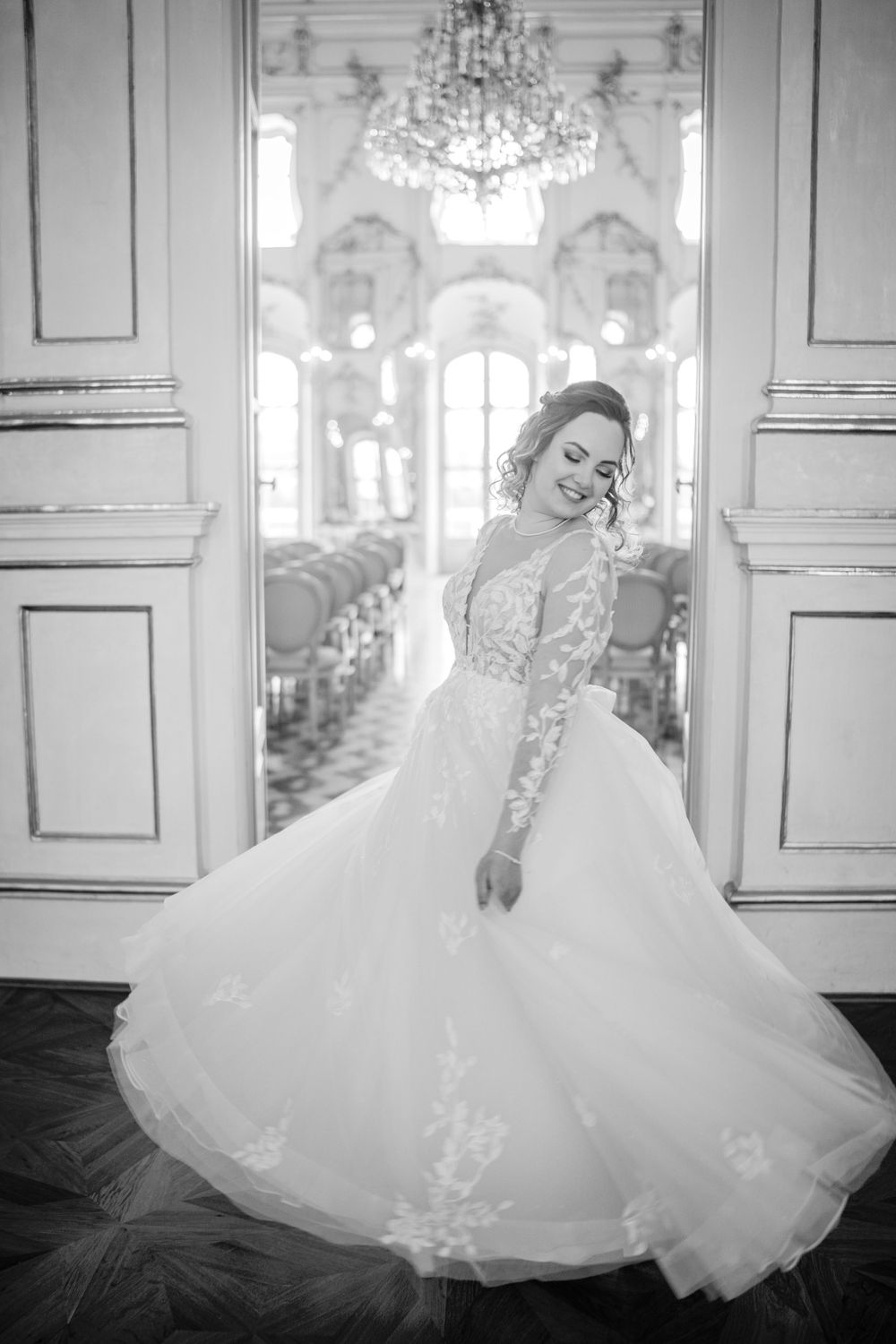 A bride in an elegant lace-sleeved wedding gown twirls in a grand ballroom with ornate chandeliers and architectural details.