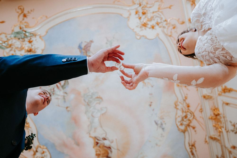 A close-up of hands exchanging wedding rings during a ceremony beneath an ornate ceiling with golden details.