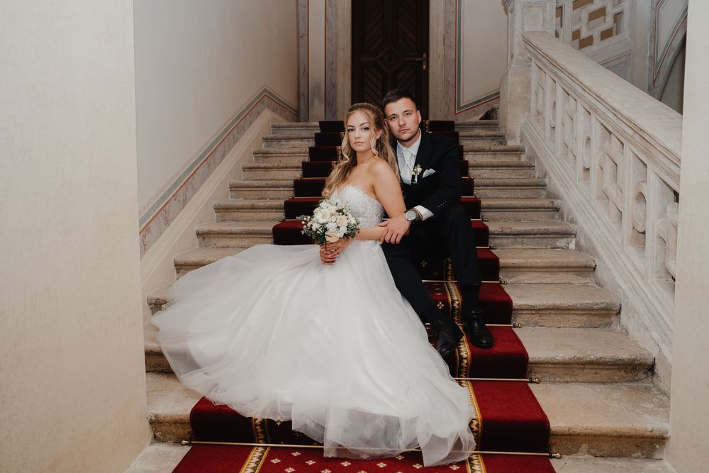 A couple sits together on grand marble stairs with a red carpet runner in an elegant venue.