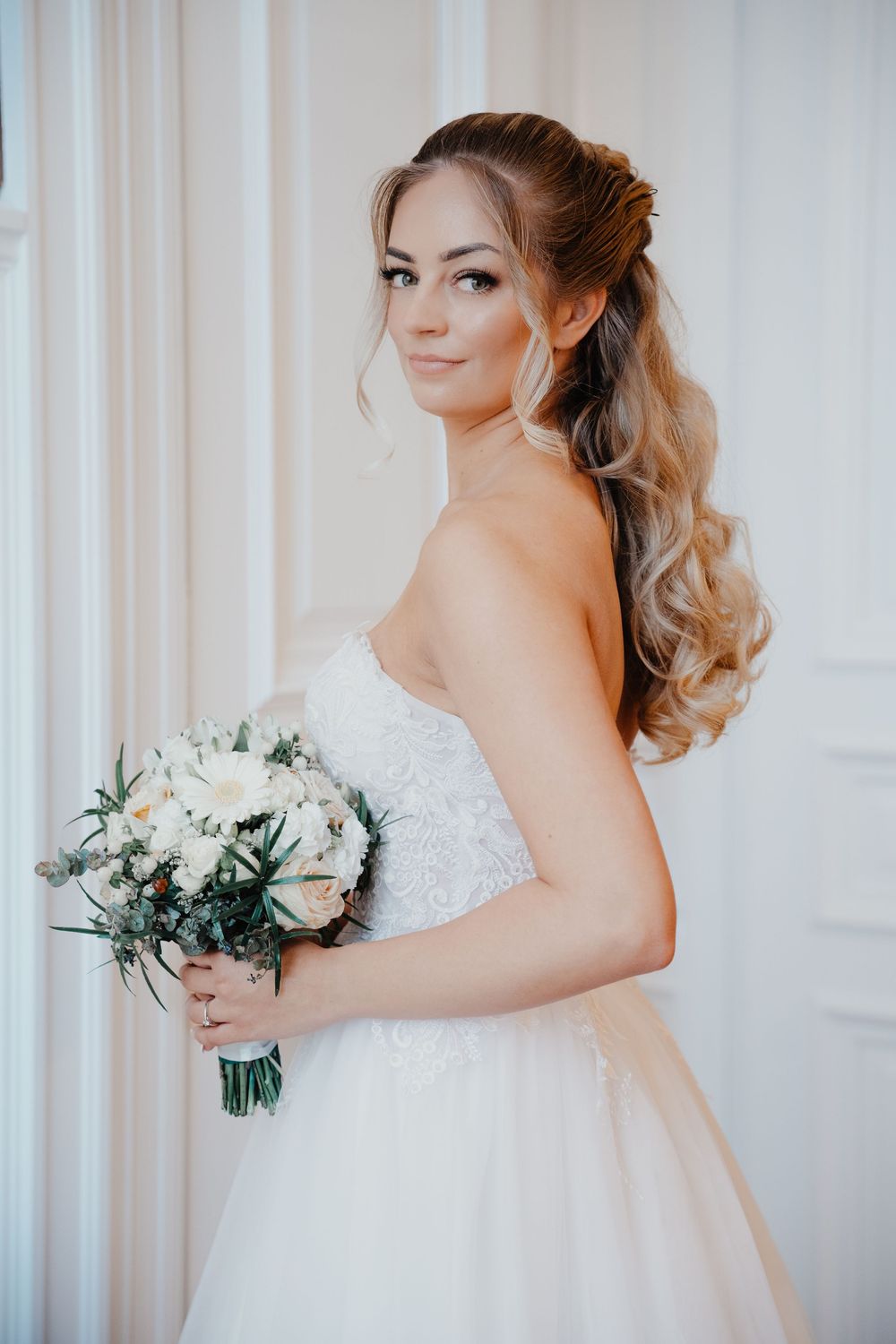 A bride in a strapless white wedding gown holds a white flower bouquet while posing against a light backdrop.