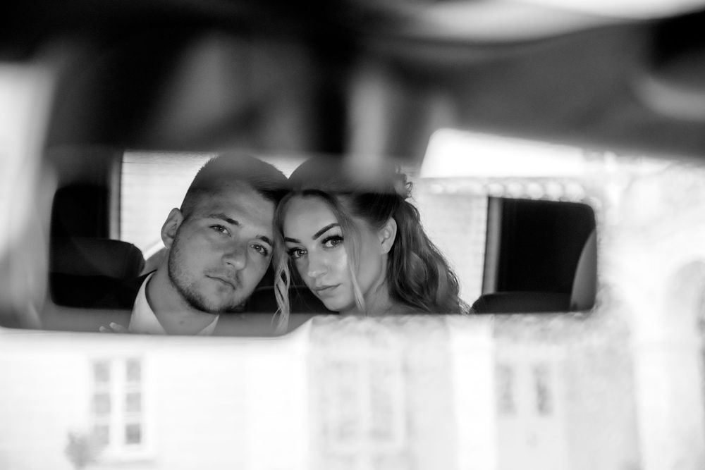 Black and white photo showing a couple's reflection in a rearview mirror of a car.