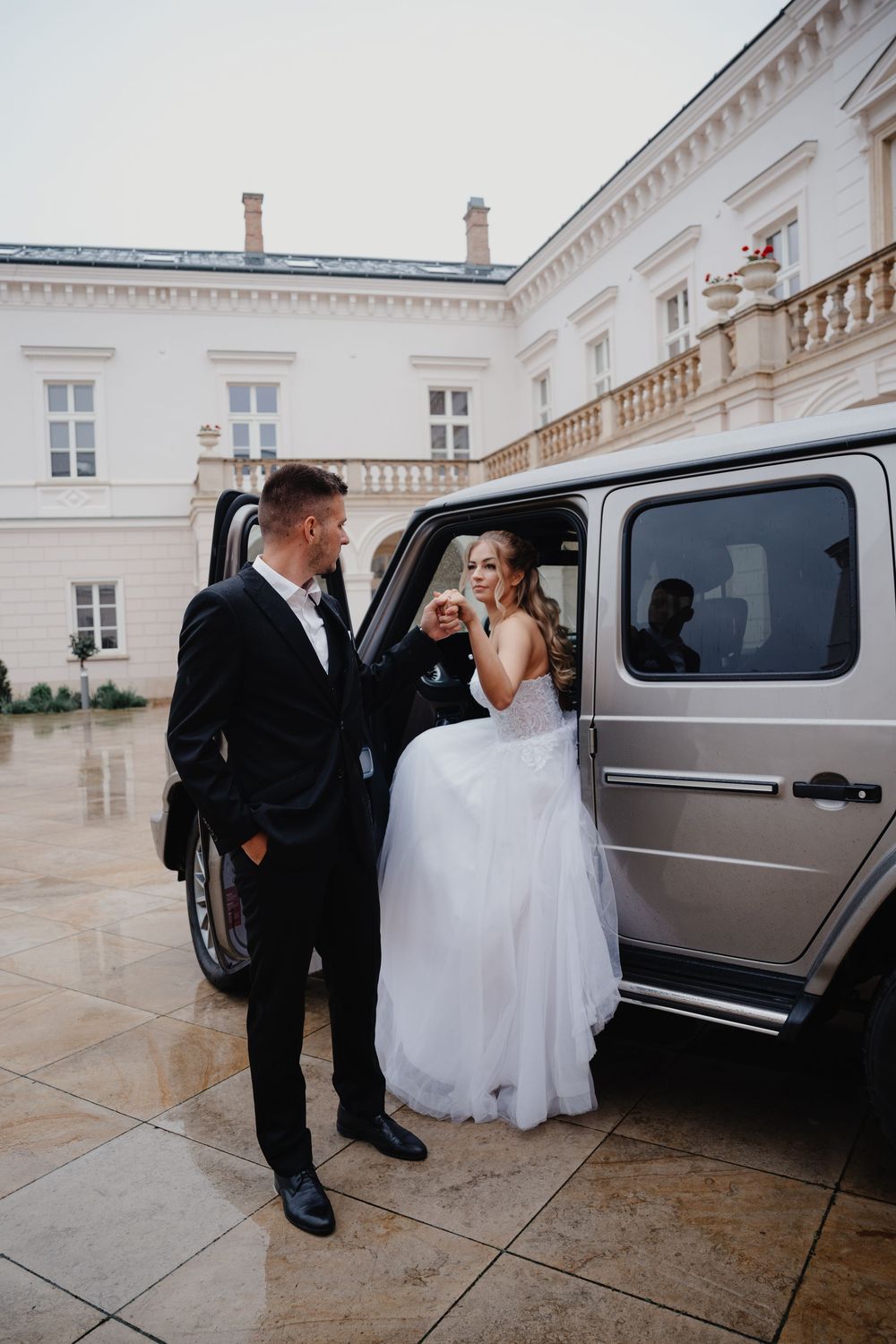 A bride in a white gown gets assistance near a luxury silver van outside an elegant historic building.