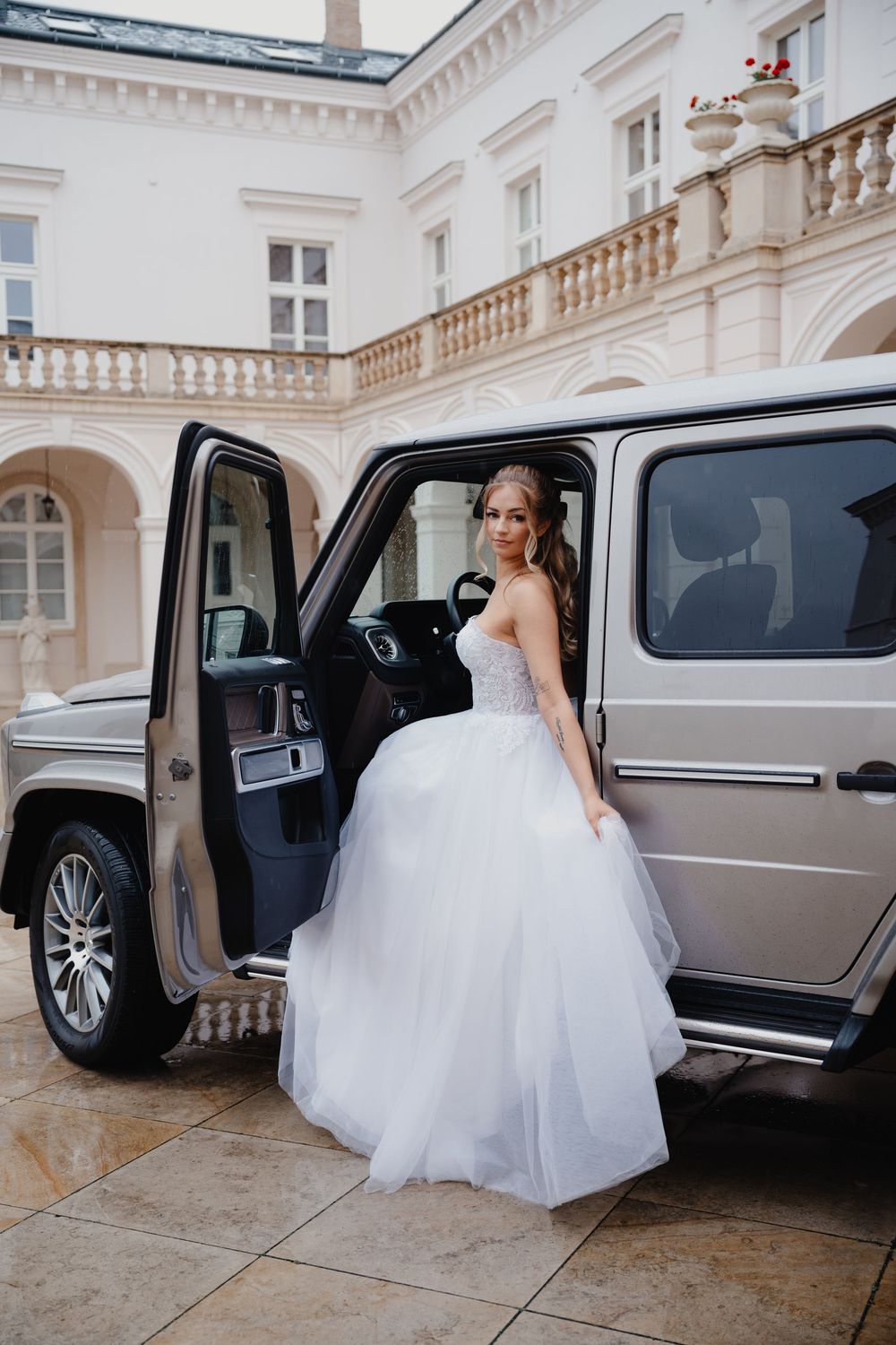 A bride in a white ballgown dress stepping out of a luxury SUV outside an elegant classical building.