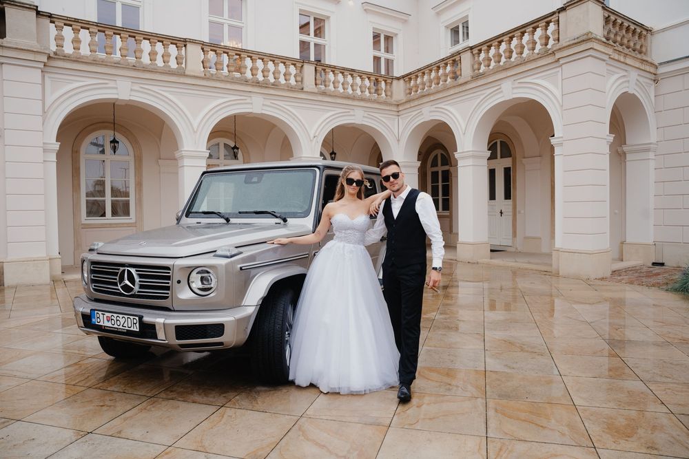A silver Mercedes G-Wagon parked in front of a grand classical building with arched columns and stone balustrades.