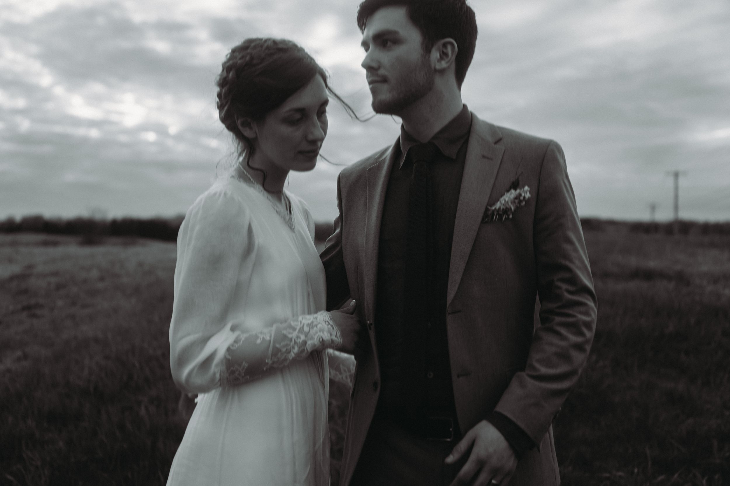 A moody black and white portrait of a couple in wedding attire standing in a field.
