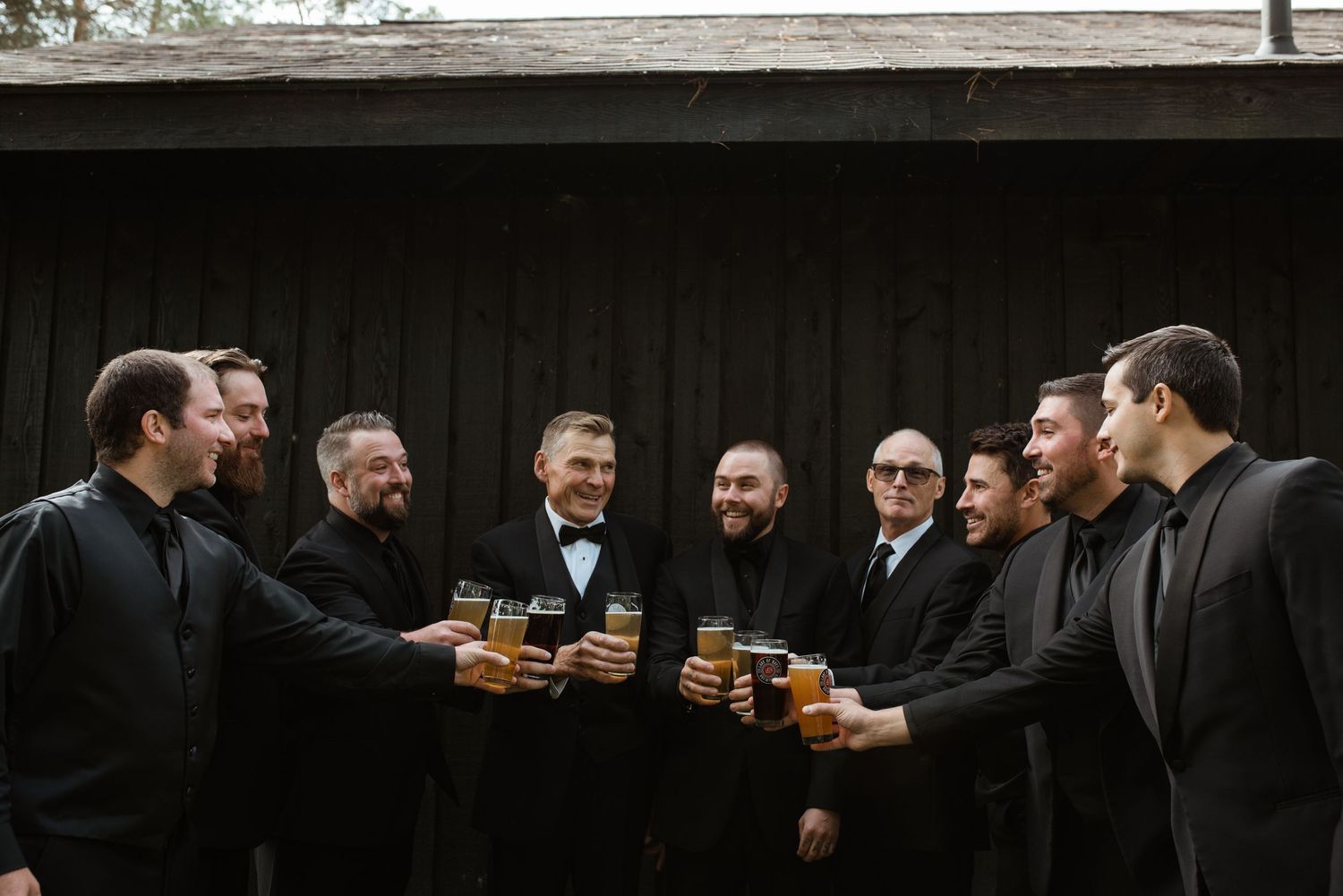 Groomsmen in dark suits toasting with beer glasses in front of a rustic wooden building.